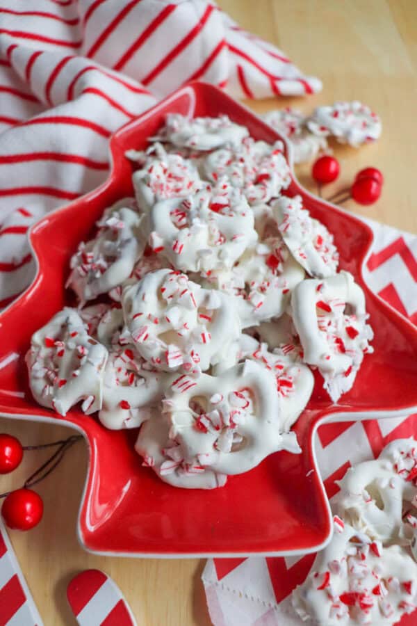 A red tree-shaped plate filled with white chocolate-coated pretzels, topped with crushed candy canes. The background features red and white striped fabric and decorative red berries, creating a festive holiday atmosphere.
