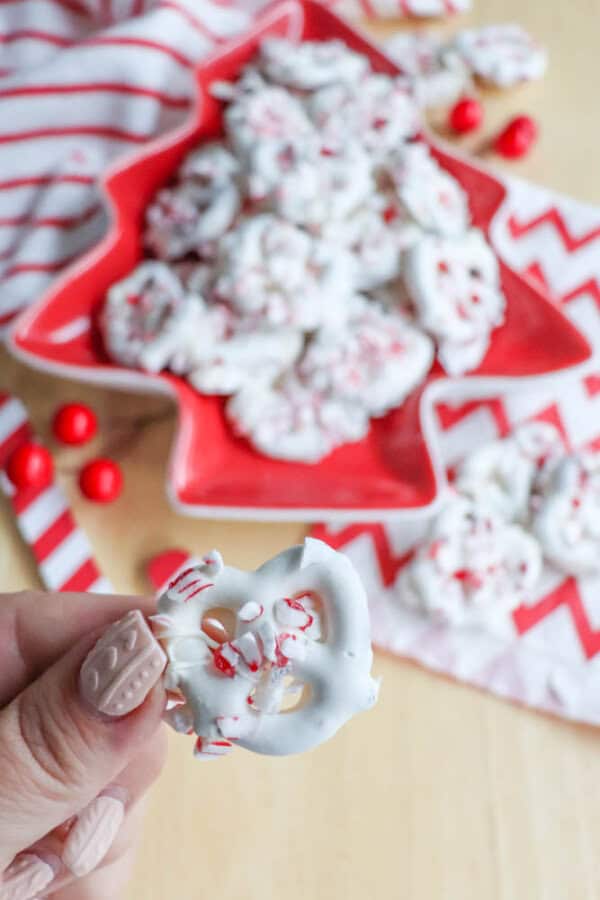 A hand holds a white chocolate-covered pretzel with crushed candy canes. In the background, more pretzels are arranged in a star-shaped red dish, surrounded by a striped cloth and red candies.