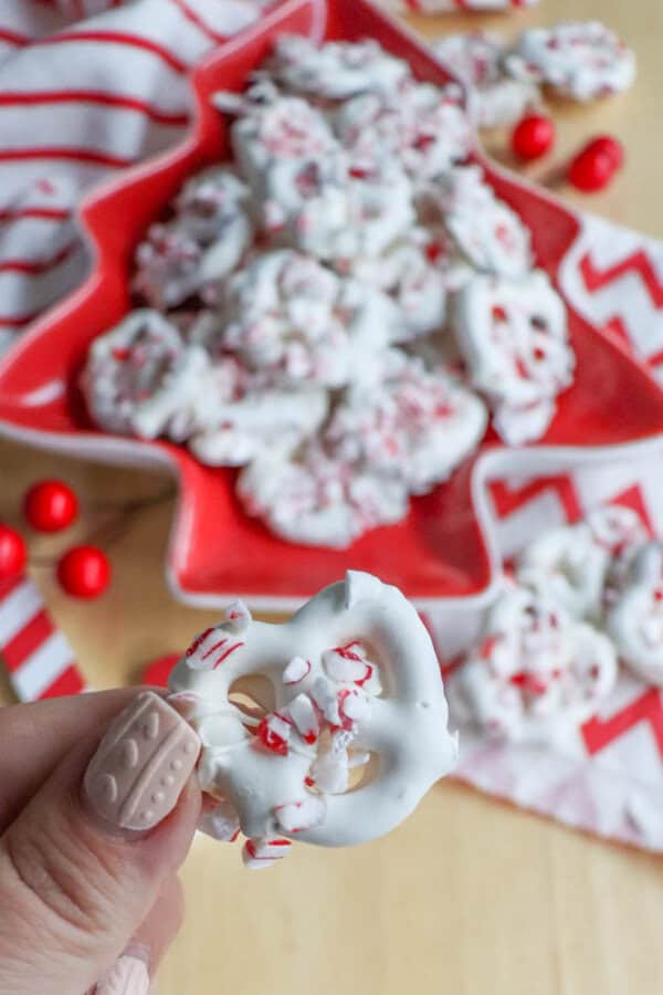 A hand holding a white chocolate-covered pretzel topped with crushed candy canes. In the background, more pretzels are arranged on a red star-shaped plate, with a striped towel and red decorations nearby.