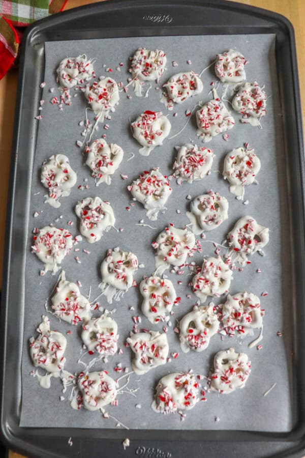 A baking tray with 24 white chocolate-covered pretzels topped with crushed candy canes, arranged on parchment paper.