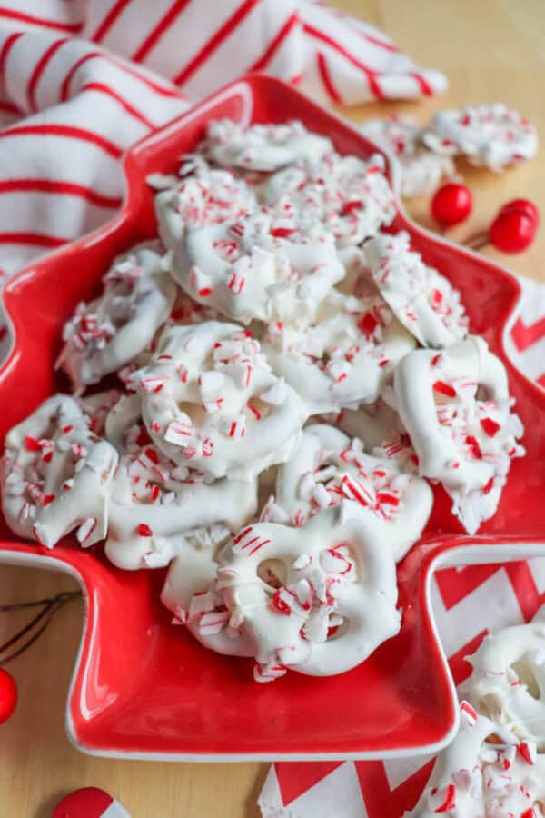 A red Christmas tree-shaped plate filled with white chocolate-covered pretzels, sprinkled with crushed peppermint candy. A red and white striped cloth and a few red candies are in the background, creating a festive holiday scene.
