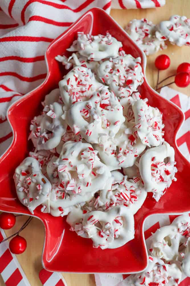 Christmas-themed plate with white chocolate-covered pretzels sprinkled with crushed peppermint. The plate is shaped like a Christmas tree, and there are festive red and white striped decorations in the background.