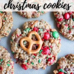 An assortment of Christmas Kitchen Sink Cookies topped with pretzels and colorful candies sits on a baking sheet. Text reads KITCHEN SINK CHRISTMAS COOKIES, followed by a website name at the bottom.