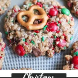 A close-up of Christmas kitchen sink cookies topped with colorful candy, chocolate chunks, and a pretzel on a baking tray. Text at the bottom reads Christmas Kitchen Sink Cookies Homemade Heather.