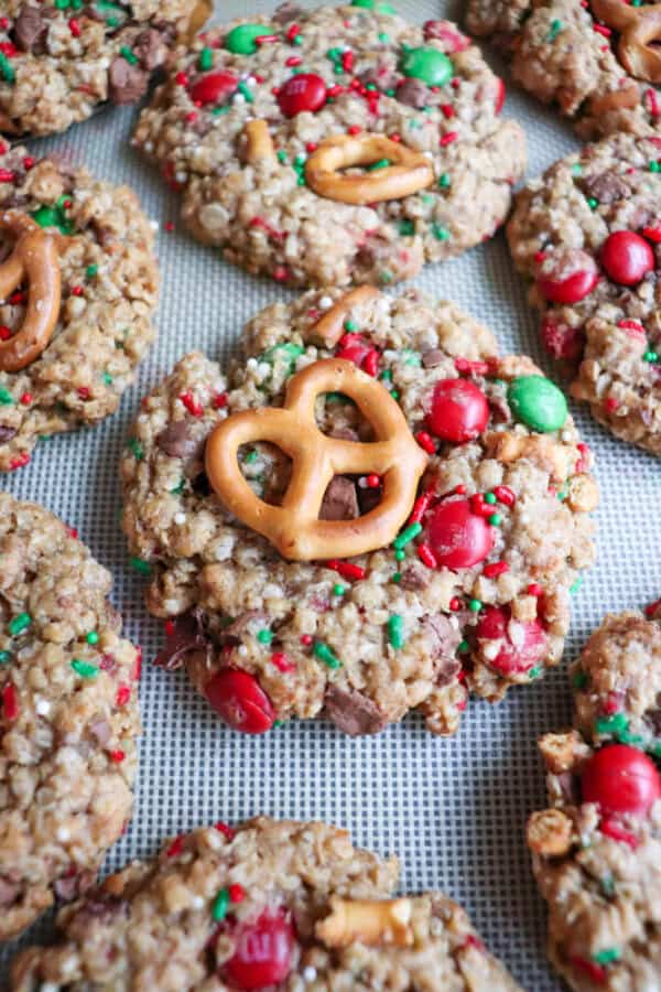 Christmas kitchen sink cookies are topped with pretzels and decorated with red and green candies and sprinkles, arranged artfully on a baking sheet. Their oatmeal or granola-like texture lends a rustic homemade appearance, perfect for festive gatherings.