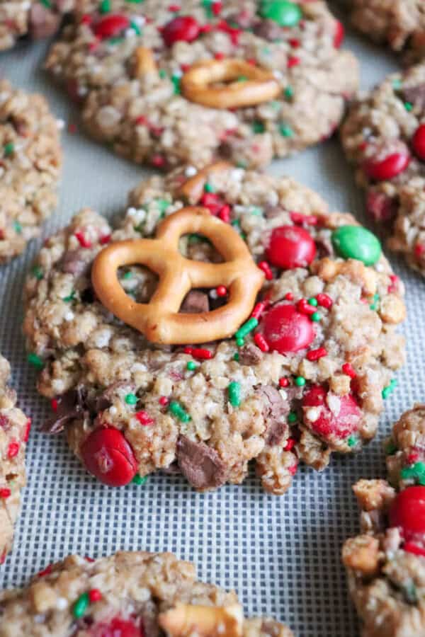 Close-up of festive Christmas kitchen sink cookies on a baking tray. Decorated with red and green candy-coated chocolates, colorful sprinkles, and topped with a pretzel, these chewy, oat-filled treats are a holiday delight.