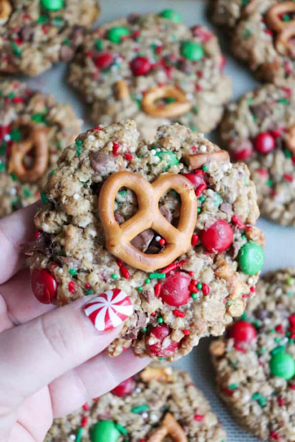 A hand with peppermint-themed nail art holds a delightful Christmas kitchen sink cookie topped with a small pretzel. The cookie is sprinkled with red and green candies and bits of chocolate. More cookies are blurred in the background, creating a festive atmosphere.