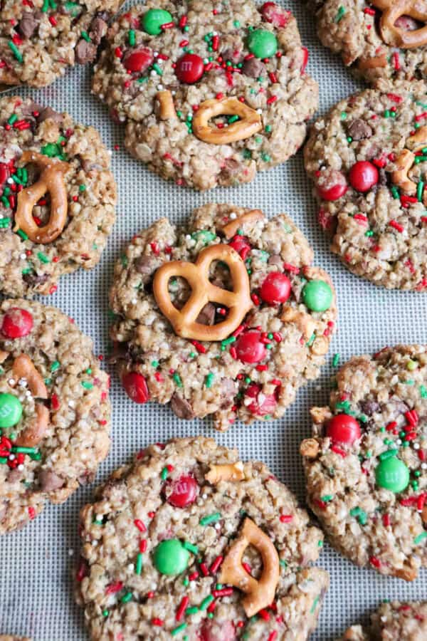 Christmas kitchen sink cookies are topped with pretzels, red and green candies, and colorful sprinkles, all carefully arranged on a baking sheet. These festive treats boast a textured, oat-filled appearance.