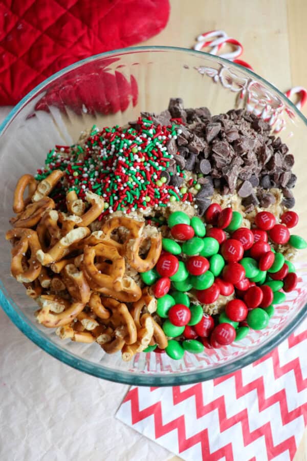 A glass bowl on a table holds chocolate chips, red and green M&Ms, colorful sprinkles, and pretzel twists—perfect ingredients for festive Christmas kitchen sink cookies. A red oven mitt and candy canes are visible in the background, while a red and white zigzag-patterned cloth rests beneath the bowl.