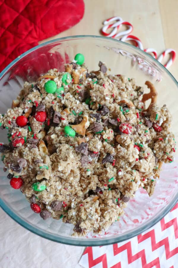 A glass bowl brims with a holiday snack mix, reminiscent of Christmas kitchen sink cookies, featuring pretzels, chocolate chips, oats, red and green candies, and sprinkles. In the background, candy canes and a red oven mitt add a festive touch on the wooden surface.
