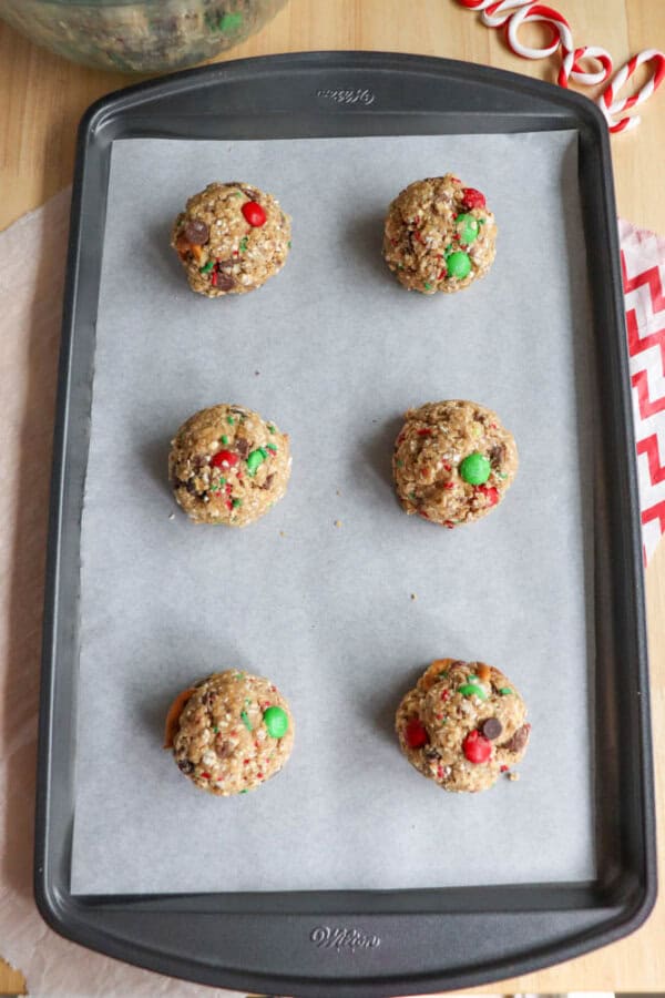 Six freshly baked Christmas cookies with red and green chocolate candies sit on a parchment-lined baking tray. A wooden surface and red-striped ribbon are partially visible in the background, creating a festive, kitchen-sink-cookie vibe.