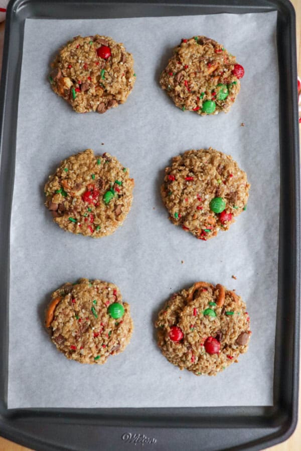 A baking sheet holds six Christmas kitchen sink cookies on parchment paper. These festive treats are dotted with red and green candies, chocolate chunks, and pretzel pieces, crafting a delightful holiday appearance.