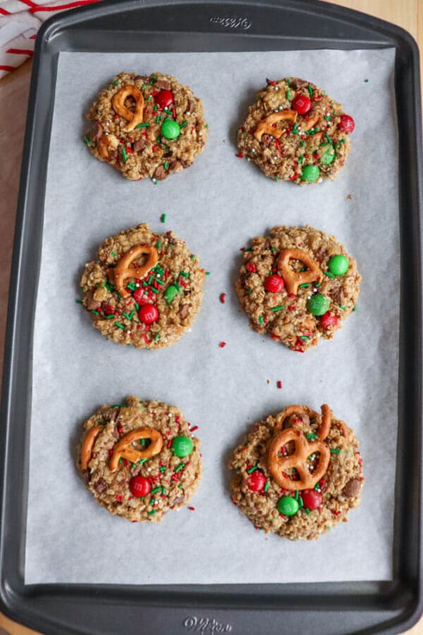 A baking tray lined with parchment paper holds six large kitchen sink cookies. Topped with pretzels, red and green candies, and colorful sprinkles, these treats capture the festive spirit of Christmas.
