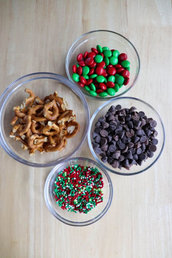Bowls on a wooden table brimming with ingredients for festive Christmas kitchen sink cookies: pretzel pieces, red and green candies, chocolate chips, and sprinkles in holiday hues of red, green, and white.