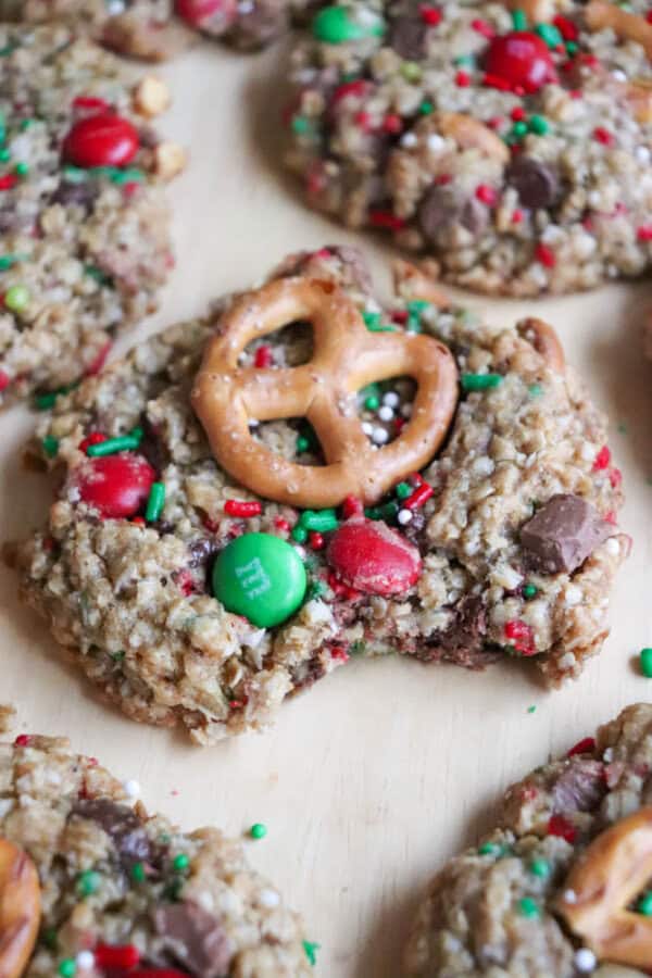 A festive Christmas cookie topped with a pretzel, red and green candies, chocolate chips, and colorful sprinkles. One bite reveals its chewy texture. Other similar kitchen sink cookies are visible in the background, making for a delightful holiday treat.