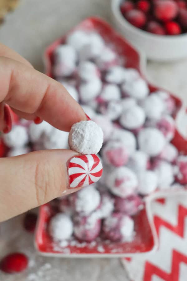A hand with peppermint nail art holds a small white powdered snack. In the background, a red tree-shaped dish is filled with similar treats and red-coated ones, resembling a festive cranberry candy recipe. A bowl of cranberries is nearby.