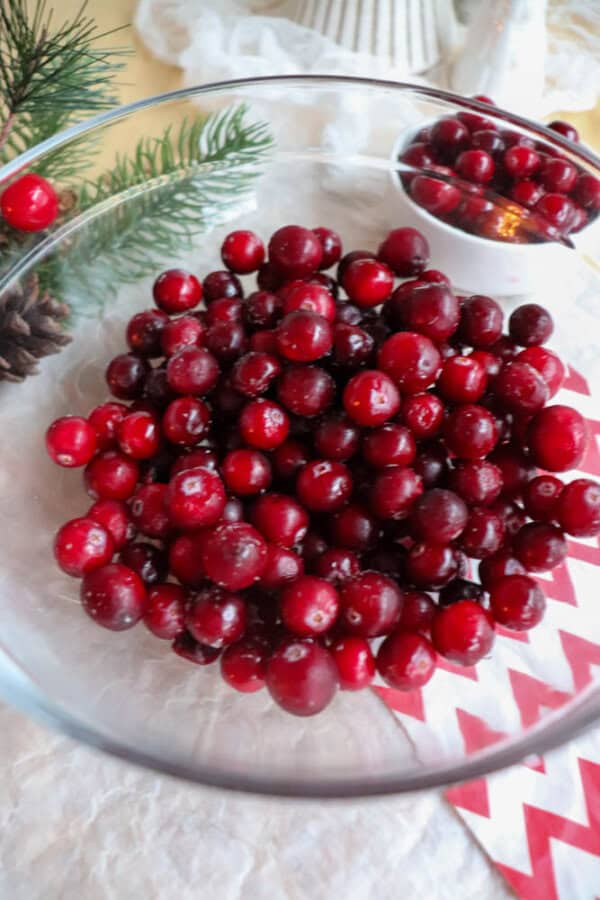 A glass bowl brimming with fresh red cranberries sits on a table adorned with a decorative pine branch, inspiring thoughts of a delightful cranberry candy recipe. In the background, a small bowl of cranberries rests beside a red and white zigzag-patterned napkin.