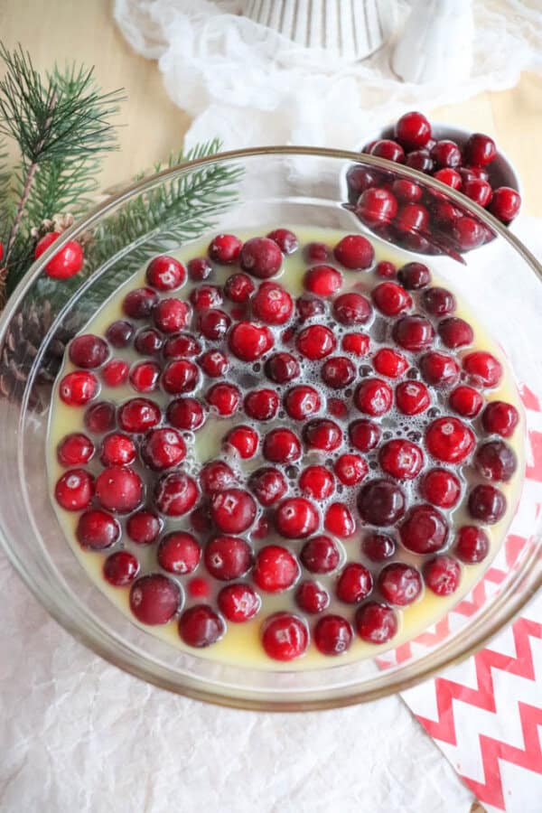 A glass bowl filled with fresh cranberries submerged in a yellow liquid is set on a table alongside a small pine branch and a chevron-patterned napkin, hinting at holiday warmth. In the background, another bowl of cranberries offers inspiration for your next cranberry candy recipe.