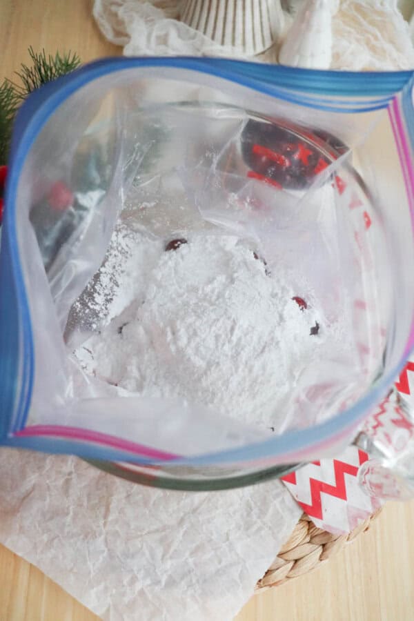 An open plastic zipper bag filled with powdered ingredients and dark pieces, perhaps chocolate or berries, hints at a cranberry candy recipe. The bag rests on crumpled parchment paper, surrounded by festive decorations adorned with red and white patterns.
