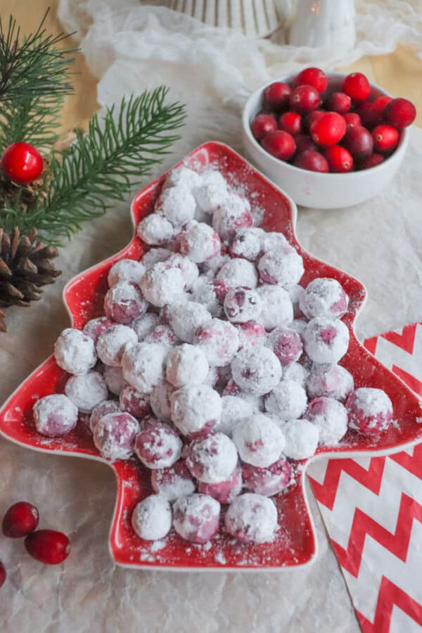A festive, tree-shaped red platter showcases sugared cranberries dusted with powdered sugar, reminiscent of a delightful cranberry candy recipe. Nearby, a white bowl brims with fresh cranberries, placed on a white cloth adorned with a pine branch and vibrant red zigzag napkins.