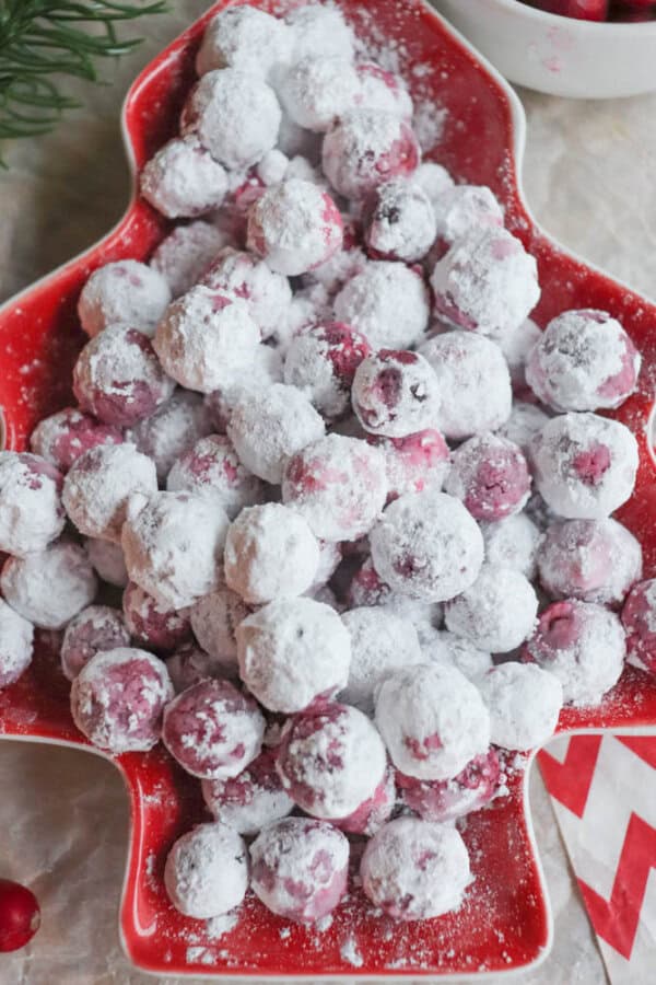A red, tree-shaped plate brimming with sugared cranberries coated in powdered sugar showcases a delightful cranberry candy recipe. A small bowl of fresh cranberries and some greenery are partially visible in the background, adding a festive touch.