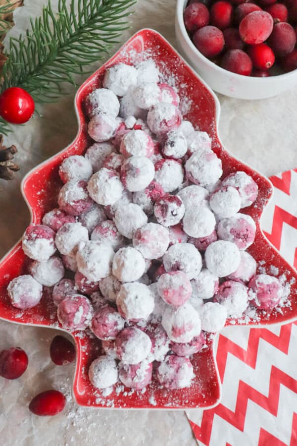 A festive Christmas tree-shaped dish brimming with sugared cranberries takes center stage on the table, a nod to a cherished cranberry candy recipe. Beside it sits a bowl of fresh cranberries and a red and white chevron-patterned napkin, with a pine branch peeking from the side.