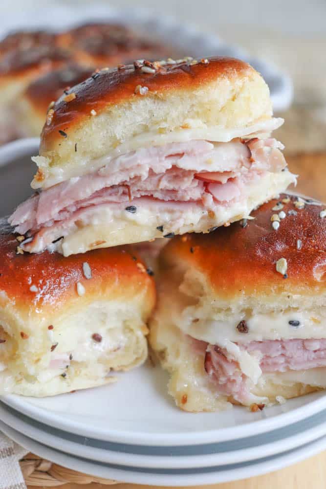 Close-up of ham and cheese sliders stacked on a plate. The sliders feature golden-brown buns with layers of ham and melted cheese, topped with a sprinkle of seasoning.