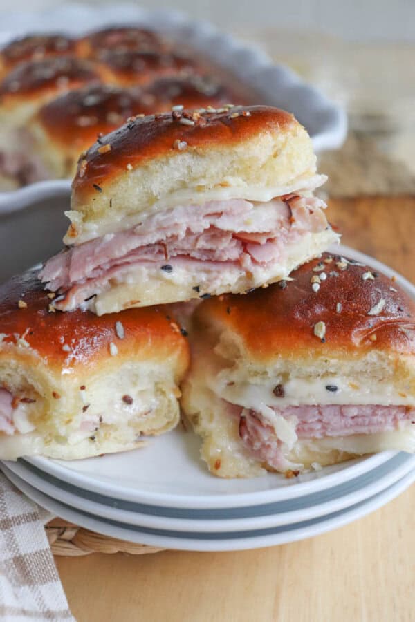 Close-up of three savory sliders stacked on a white plate. The sliders have glossy tops and are filled with ham, cheese, and a sprinkling of seeds. A dish with more sliders is blurred in the background.