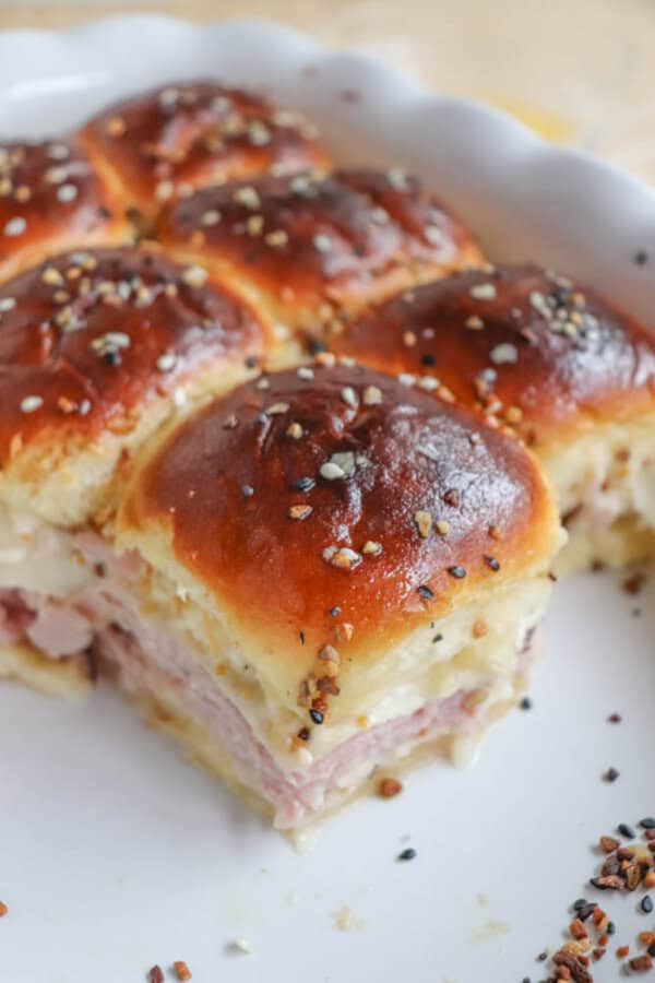A close-up of baked sliders on a white plate. The sliders have a golden-brown top sprinkled with sesame seeds and spices. Layers of ham and melted cheese are visible between the soft buns. One slider is partially removed, revealing a side view.