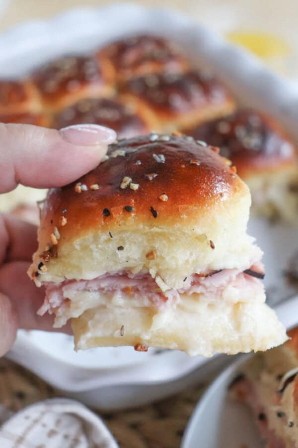 A hand holding a small sandwich made with a shiny, golden-brown roll filled with layers of ham and melted cheese. In the background, more sandwiches are visible on a white baking dish. The roll is sprinkled with seeds and seasoning.