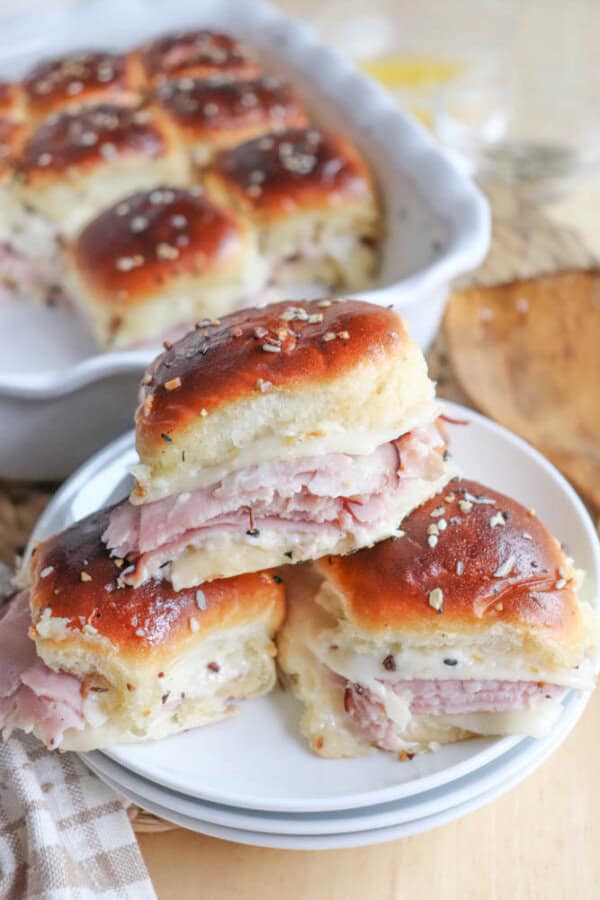 A close-up of three ham and cheese sliders stacked on a white plate. The sliders have golden brown tops sprinkled with sesame seeds. In the background, a baking dish with more sliders is visible.