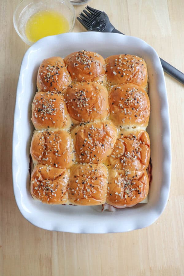 A white baking dish filled with twelve freshly baked slider rolls topped with sesame and poppy seeds. A bowl of melted butter and a basting brush are placed nearby on a wooden surface.