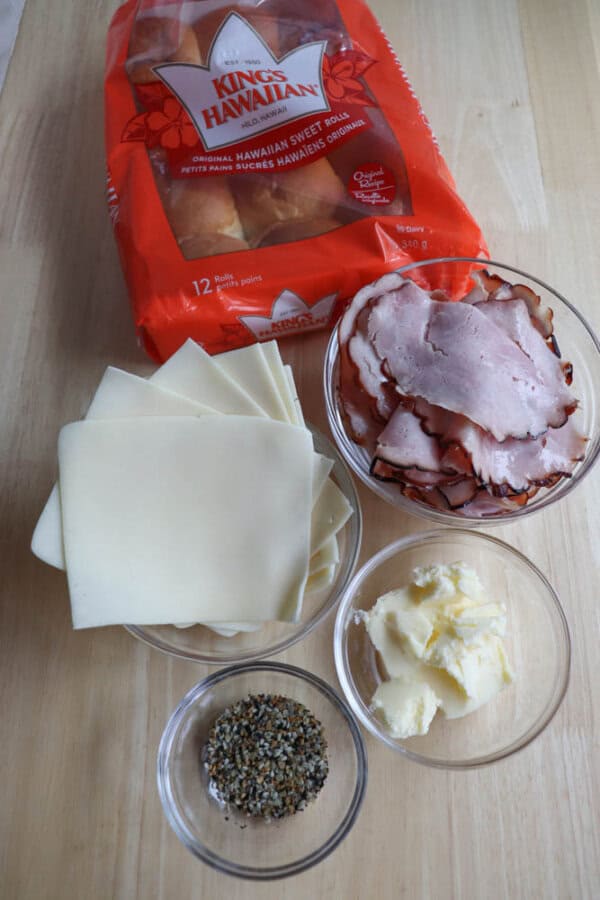 Ingredients on a table include a package of Kingโs Hawaiian rolls, slices of cheese, deli meat, a bowl of butter, and a bowl of seasoning.