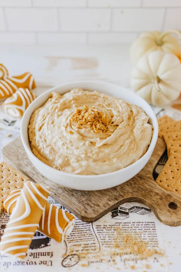 A bowl of creamy Little Debbie pumpkin spice dip garnished with crushed biscuits sits on a wooden board. Surrounding the bowl are graham crackers and wrapped caramel candies. In the background, there's a white pumpkin and a newspaper.