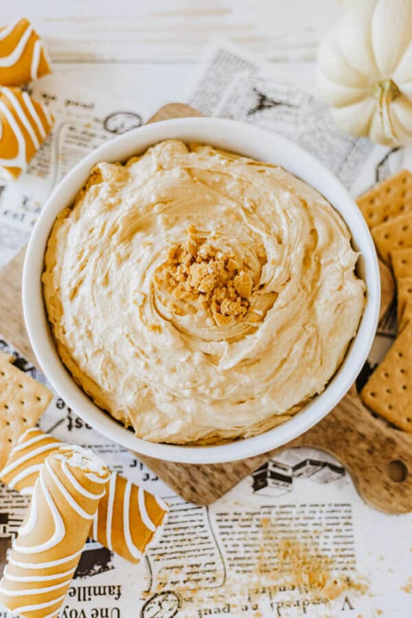 A bowl of creamy pumpkin dip, reminiscent of Little Debbie's pumpkin spice delight, is topped with crumbled crackers and surrounded by whole crackers and small white pumpkins. The setup rests on a wooden board over a newspaper, with swirled orange and white napkins elegantly flanking the arrangement.