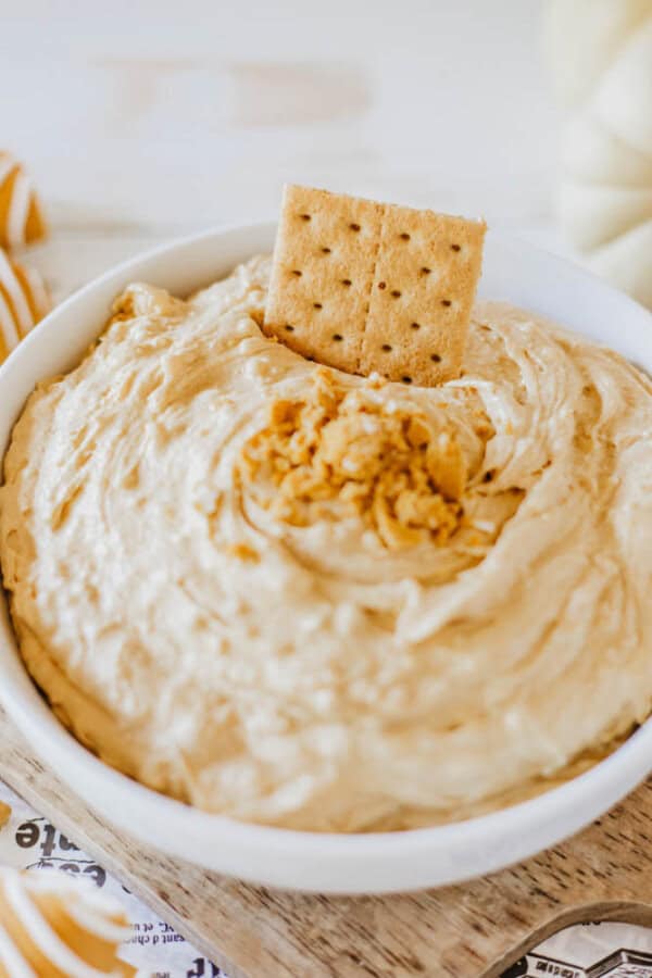 A white bowl filled with creamy Little Debbie Pumpkin Spice dip, topped with breadcrumbs and garnished with a rectangular cracker, rests on a wooden board with a striped cloth nearby.