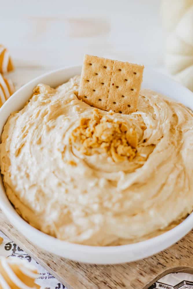 A white bowl filled with creamy Little Debbie Pumpkin Spice dip, topped with breadcrumbs and garnished with a rectangular cracker, rests on a wooden board with a striped cloth nearby.