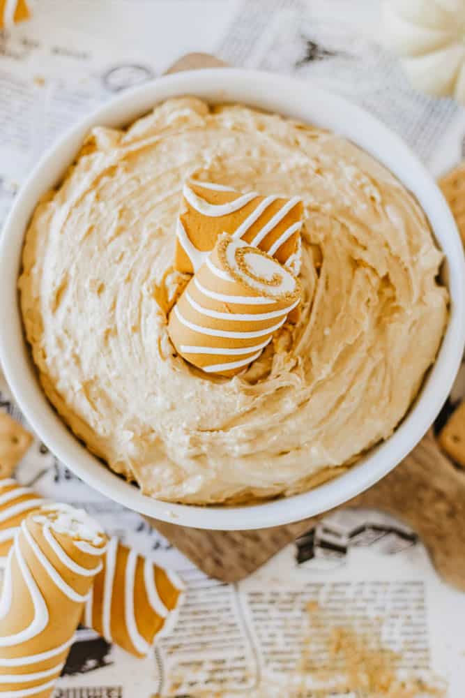 A bowl filled with creamy, light brown Little Debbie pumpkin spice cheesecake dip. Two rolled wafers with white icing stand upright in the dip, surrounded by scattered wafers on a table with printed graphics.