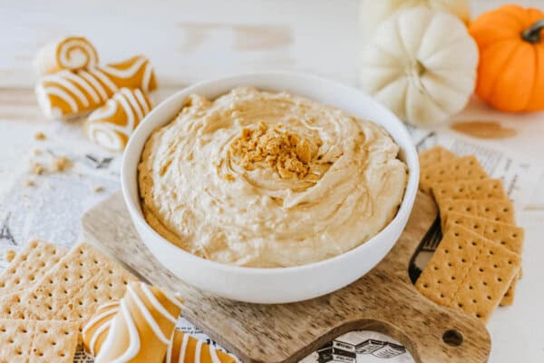 A bowl of creamy Little Debbie Pumpkin Spice dip topped with crumbles sits on a wooden board surrounded by graham crackers. In the background, there are white and orange mini pumpkins and striped cookies. The scene has an autumnal feel.