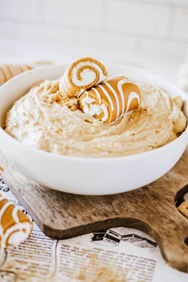 A white bowl filled with creamy Little Debbie pumpkin spice dip, topped with two rolled cookies drizzled with white icing. The bowl is nestled on a wooden board, with a newspaper partially visible underneath.