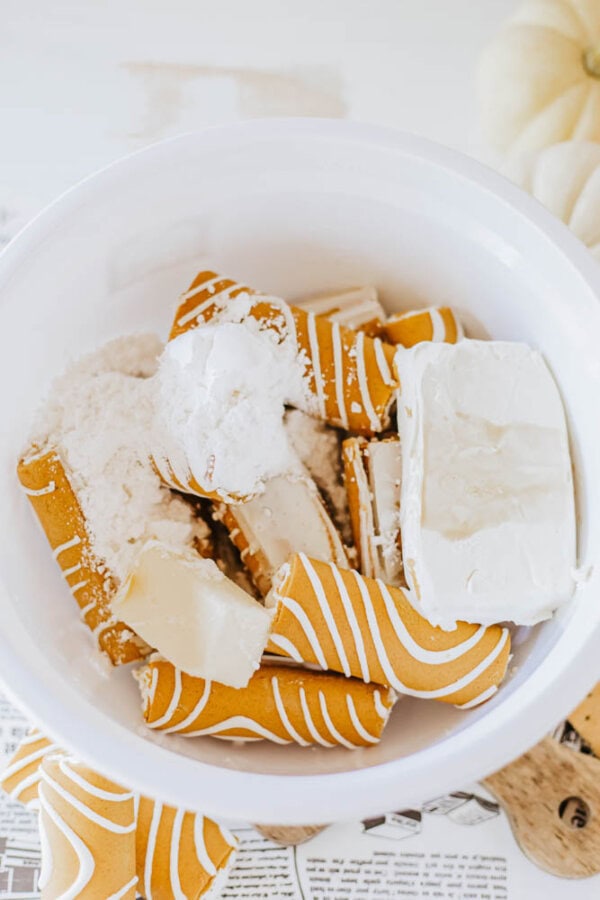 A white mixing bowl holds broken pumpkin spice wafer rolls, cream cheese, and powdered sugar, ready to become a delightful Little Debbie pumpkin spice dip. A small pumpkin peeks into view from the background, hinting at the seasonal flavor awaiting to be mixed.