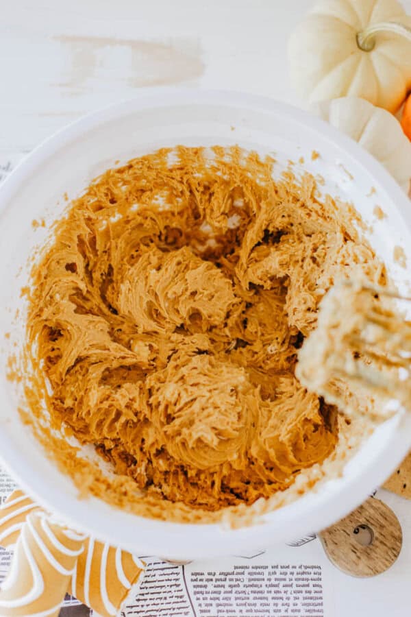 A bowl of pumpkin cookie dough sits on a table, next to a mixer. Nearby are small pumpkins and pumpkin-shaped cookies with orange icing. A hint of Little Debbie pumpkin spice dip adds to the setting's cozy, autumnal feel.