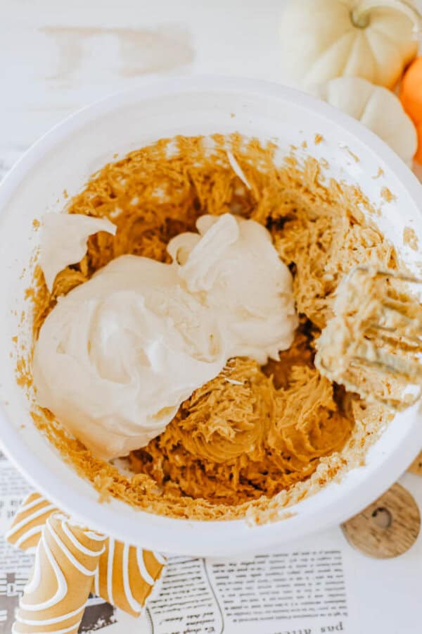 A bowl of cookie dough is being mixed, with a creamy ingredient, possibly Little Debbie pumpkin spice dip, partially blended in. The mixture is a light brown color. A striped cloth and part of a page are on the table nearby, alongside a small pumpkin decoration.