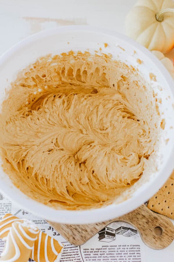 A bowl of creamy, light brown cookie dough sits on a table surrounded by a small pumpkin, graham crackers, and a decorative frosted pumpkin-shaped cookie. The swirl of the Little Debbie Pumpkin Spice Dip adds smooth excitement, ready to enhance any baking adventure.