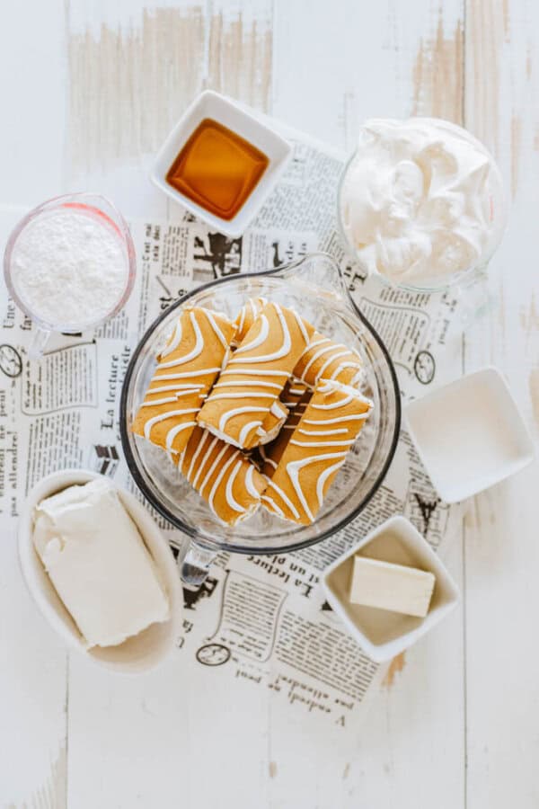 Ingredients for a dessert on a table, featuring Little Debbie pumpkin spice dip, a bowl of striped cookies, a glass of whipped cream, white icing, cream cheese, butter, and vanilla extract—all artfully laid out on a newspaper.