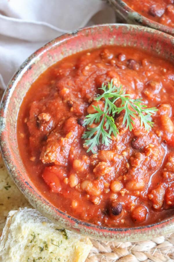 A bowl of hearty chili with beans, tomato sauce, and ground meat, made as an easy crockpot chili, is garnished with a sprig of fresh parsley. A slice of garlic bread is placed nearby.