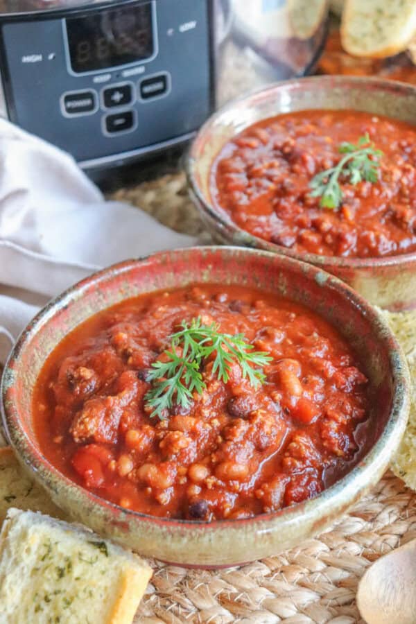 Two bowls of hearty, easy crockpot chili topped with fresh parsley sit on a woven mat. Slices of bread are nearby, and a slow cooker is in the background. A wooden spoon rests beside the bowls, adding to the cozy, homemade feel.