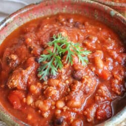 A bowl of hearty, easy crockpot chili topped with a sprig of fresh parsley. The chili is rich with beans, tomatoes, and ground meat, creating a thick, savory delight. A piece of bread is partially visible at the bottom for dipping pleasure.