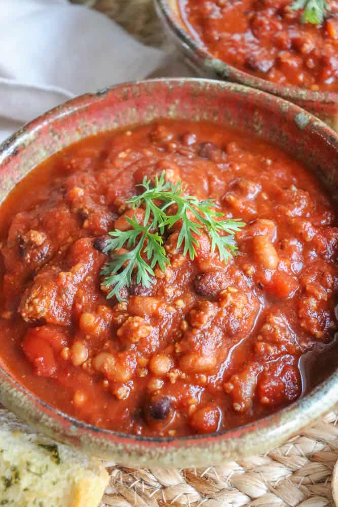 A bowl of hearty, easy crockpot chili topped with a sprig of fresh parsley. The chili is rich with beans, tomatoes, and ground meat, creating a thick, savory delight. A piece of bread is partially visible at the bottom for dipping pleasure.