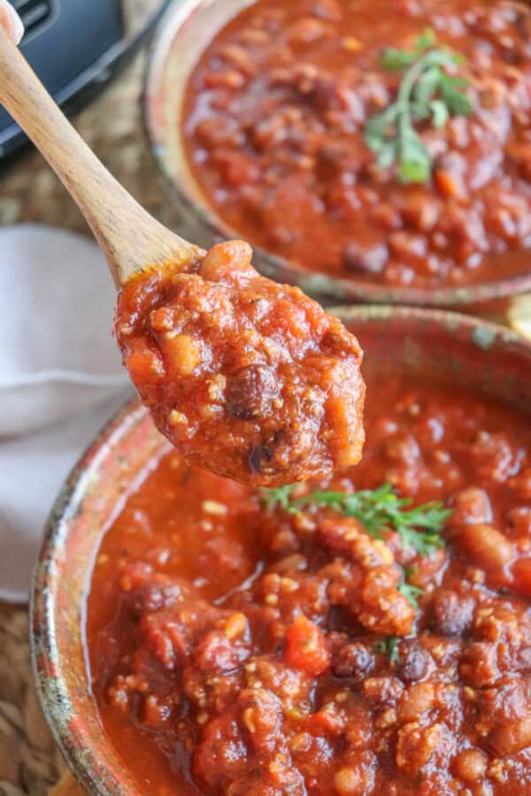 A wooden spoon holds a serving of hearty, easy crockpot chili filled with beans and meat, lifted above a bowl garnished with fresh herbs. The chili's rich, red hue hints at its robust tomato-based sauce.
